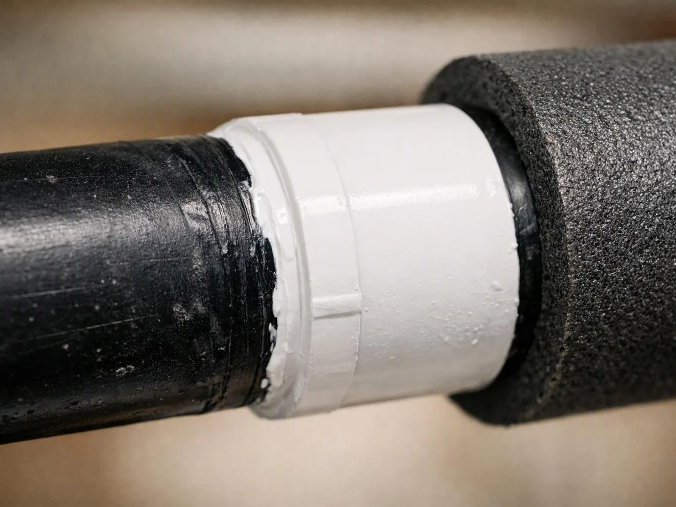 A black pipe joins a white connector wrapped in gray foam insulation against a blurred beige background